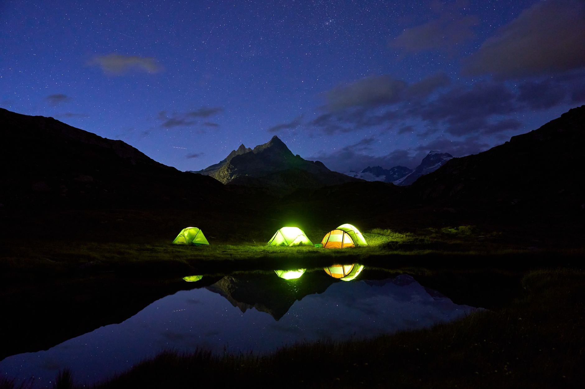 A tranquil night camping by a mountain lake with glowing tents under a starlit sky.
