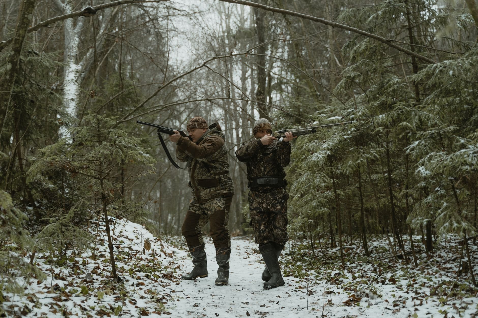 Two hunters in camouflage walking through a snowy forest with rifles.